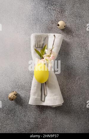 Besteck mit Frühlingsdekorationen, gelben Eiern und Blütenblumen für die Ostertafel auf grauem Hintergrund. Blick von oben. Vertikales Format. Stockfoto