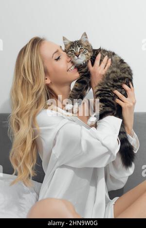 Porträt von jungen schönen lächelnden Frau mit langen welligen hellen Haaren in weißen losen Hemd kuscheln junge niedliche gestreifte Katze. Stockfoto