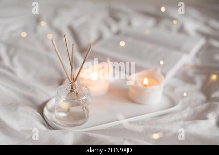 Heimisch flüssiger Duft in Glasflasche und brennende Kerzen bleiben auf weißem Keramiktablett im Bett aus nächster Nähe. Gemütliche und hygge Atmosphäre. Stockfoto