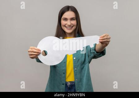 Porträt einer glücklichen dunkelhaarigen Frau, die den Papierschlüssel als Symbol für die eigene Wohnung hält, Träume werden wahr, staatliches Wohnprogramm, in lässiger Jacke. Innenaufnahme des Studios isoliert auf grauem Hintergrund. Stockfoto