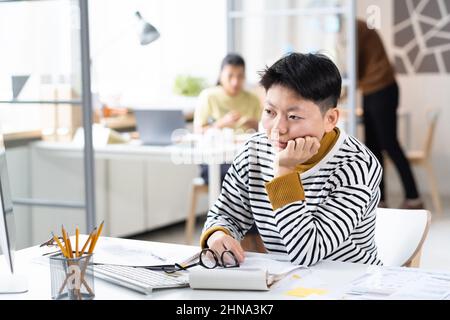Asiatische Geschäftsfrau, die an ihrem Arbeitsplatz im Büro sitzt und mit gelangweilter Ausdrucksweise auf den Computermonitor schaut Stockfoto