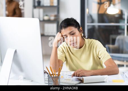 Junge Geschäftsfrau, die mit einem Computermonitor an ihrem Arbeitsplatz sitzt und über etwas nachdenkt, ist ihrer Arbeit müde Stockfoto