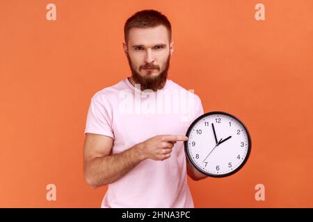 Porträt eines ernsthaften bärtigen Mannes, der mit dem Finger auf eine große Wanduhr zeigt und die Kamera anschaut, besorgt über die zeitgerecht abgeschlossene Arbeit, trägt ein rosa T-Shirt. Innenaufnahme des Studios isoliert auf orangefarbenem Hintergrund. Stockfoto