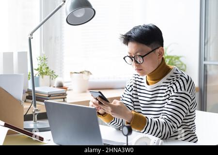 Asiatische Geschäftsfrau in Brillen sitzt an ihrem Arbeitsplatz mit Laptop und tippt eine Nachricht auf ihr Handy Stockfoto