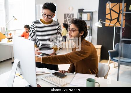 Ein junger Geschäftsmann, der am Tisch sitzt und auf einen Computermonitor zeigt, diskutierte während der Arbeit im Büro mit seinem Kollegen über das Online-Dokument Stockfoto