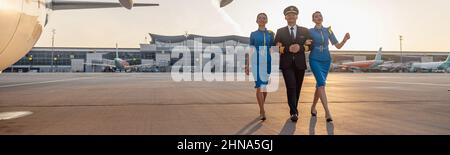 In voller Länge Aufnahme eines aufgeregten männlichen Piloten, der zusammen mit zwei weiblichen Flugbegleitern in blauer Uniform vor einem Flugzeug im Terminal bei läuft Stockfoto