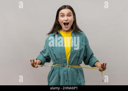 Porträt einer zufriedenen, glücklichen, schlanken Frau, die sich am Taillenband hält und die Kamera mit Spannung anschaut und eine Jacke im lässigen Stil trägt. Innenaufnahme des Studios isoliert auf grauem Hintergrund. Stockfoto