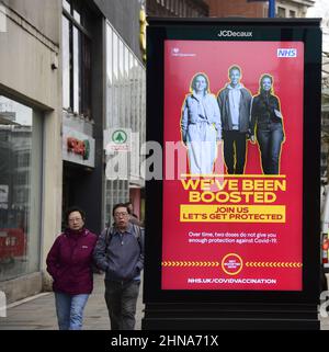 Die Menschen laufen an einem elektronischen Schild der Regierung NHS und HM vorbei, das „Wir wurden aufgestockt“ wurde, um für den Auffrischungsimpfstoff zur Bekämpfung von Covid-19 in Manchester, Greater Manchester, England, Großbritannien, zu werben. Die Zahl der Covid-19-, Coronavirus- oder Corona-Infektionen im Großraum Manchester sinkt weiter. In der Woche bis zum 8th. Februar 2022 wurden in der Region 12.364 Personen positiv getestet, was einem Rückgang um 35 Prozent gegenüber der Vorwoche entspricht. Stockfoto