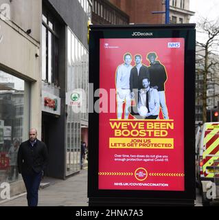 Die Menschen laufen an einem elektronischen Schild der Regierung NHS und HM vorbei, das „Wir wurden aufgestockt“ wurde, um für den Auffrischungsimpfstoff zur Bekämpfung von Covid-19 in Manchester, Greater Manchester, England, Großbritannien, zu werben. Die Zahl der Covid-19-, Coronavirus- oder Corona-Infektionen im Großraum Manchester sinkt weiter. In der Woche bis zum 8th. Februar 2022 wurden in der Region 12.364 Personen positiv getestet, was einem Rückgang um 35 Prozent gegenüber der Vorwoche entspricht. Stockfoto