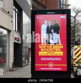 Die Menschen laufen an einem elektronischen Schild der Regierung NHS und HM vorbei, das „Wir wurden aufgestockt“ wurde, um für den Auffrischungsimpfstoff zur Bekämpfung von Covid-19 in Manchester, Greater Manchester, England, Großbritannien, zu werben. Die Zahl der Covid-19-, Coronavirus- oder Corona-Infektionen im Großraum Manchester sinkt weiter. In der Woche bis zum 8th. Februar 2022 wurden in der Region 12.364 Personen positiv getestet, was einem Rückgang um 35 Prozent gegenüber der Vorwoche entspricht. Stockfoto