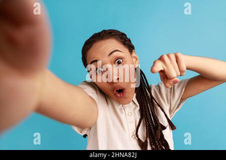 Schockierte Frau mit schwarzen Dreadlocks hat erstaunt Gesichtsausdruck, macht Selfie, drückt überrascht Emotionen, zeigt Finger auf Kamera, POV. Innenaufnahme des Studios isoliert auf blauem Hintergrund. Stockfoto