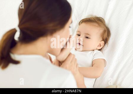 Glückliche, liebevolle Mutter küsst die kleinen Füße ihres kleinen Jungen, während sie zu Hause auf dem Bett kuschelt Stockfoto