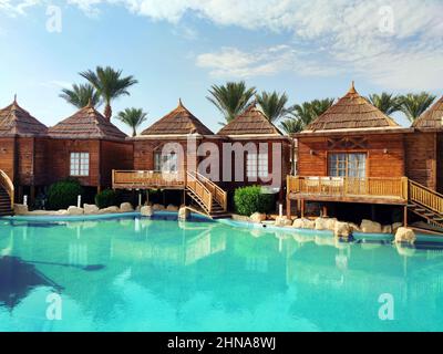 Kleine Häuser mit Strohdach in der Nähe eines Pools mit blauem Wasser, gegen einen bewölkten Himmel und grüne Palmen Stockfoto