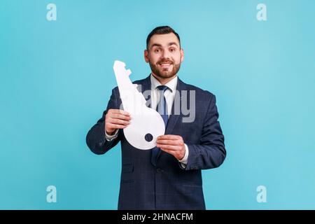 Porträt eines Mannes, der einen offiziellen Anzug trägt, der einen großen Papierschlüssel in der Hand hält, als Symbol für ihre eigene Wohnung, Träume werden wahr, staatliches Wohnungsprogramm. Innenaufnahme des Studios isoliert auf blauem Hintergrund. Stockfoto