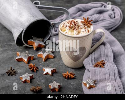 Eine Tasse gefüllt mit Schlagsahne und Kakaopulver ist mit Sternanis, Kokosnusssternen, einem grauen Schal und einer Vintage-Vase auf dem Tisch dekoriert Stockfoto