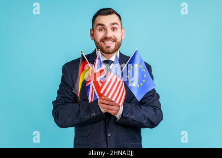 Positiver Mann mit Bart, der einen Anzug im offiziellen Stil trägt, der Flaggen verschiedener Länder hält, Sprachen lernt, die Kamera mit einem toothy Lächeln anschaut. Innenaufnahme des Studios isoliert auf blauem Hintergrund. Stockfoto