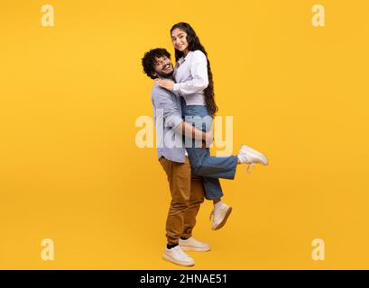 Romantischer junger inder, der seine Freundin hebt und sie hält, das Paar schaut und lächelt auf die Kamera, gelber Hintergrund Stockfoto