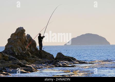 Benidorm Alicante Spain 02.15.22 Übersicht eines älteren Mannes mit Mütze, der auf einem zerklüfteten Felsvorsprung am Meer steht, der sich in einer Angelrutenschnur schlängelt. Ruhiges Blau Stockfoto