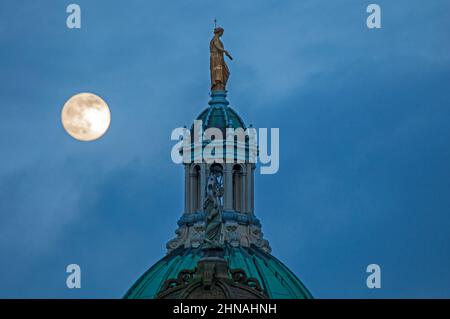 Edinburgh, Schottland, Großbritannien. 15th. Februar 2022. Der Schneemond, der morgen zu 100 % voll sein wird, erscheint am Himmel hinter der Statue, die Fame auf der zentralen achteckigen Kuppel des ehemaligen Hauptquartiers der Bank of Scotland auf dem Hügel darstellt. Wetter sehr bewölkt mit gelegentlichen Wolkenlücken und Temperaturen von 4 Grad Celsius und heftigem Regen kurz nach diesem Shooting. Stockfoto