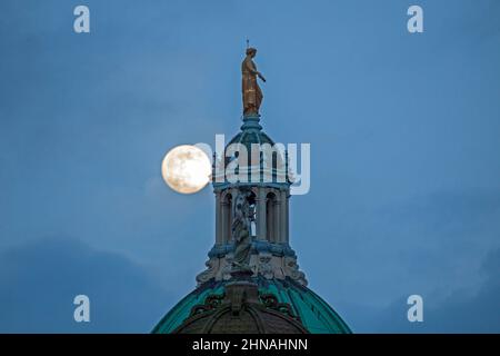 Edinburgh, Schottland, Großbritannien. 15th. Februar 2022. Der Schneemond, der morgen zu 100 % voll sein wird, erscheint am Himmel hinter der Statue, die Fame auf der zentralen achteckigen Kuppel des ehemaligen Hauptquartiers der Bank of Scotland auf dem Hügel darstellt. Wetter sehr bewölkt mit gelegentlichen Wolkenlücken und Temperaturen von 4 Grad Celsius und heftigem Regen kurz nach diesem Shooting. Stockfoto