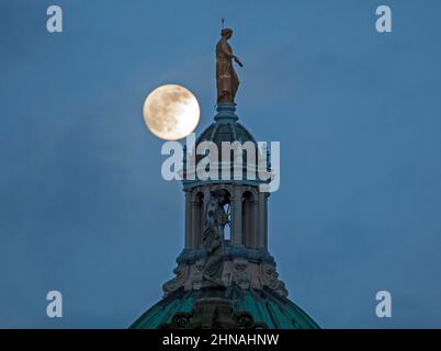 Edinburgh, Schottland, Großbritannien. 15th. Februar 2022. Der Schneemond, der morgen zu 100 % voll sein wird, erscheint am Himmel hinter der Statue, die Fame auf der zentralen achteckigen Kuppel des ehemaligen Hauptquartiers der Bank of Scotland auf dem Hügel darstellt. Wetter sehr bewölkt mit gelegentlichen Wolkenlücken und Temperaturen von 4 Grad Celsius und heftigem Regen kurz nach diesem Shooting. Stockfoto