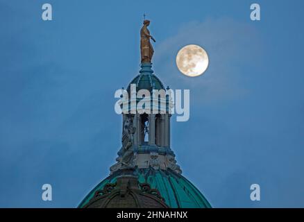 Edinburgh, Schottland, Großbritannien. 15th. Februar 2022. Der Schneemond, der morgen zu 100 % voll sein wird, erscheint am Himmel hinter der Statue, die Fame auf der zentralen achteckigen Kuppel des ehemaligen Hauptquartiers der Bank of Scotland auf dem Hügel darstellt. Wetter sehr bewölkt mit gelegentlichen Wolkenlücken und Temperaturen von 4 Grad Celsius und heftigem Regen kurz nach diesem Shooting. Stockfoto