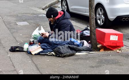 Mann, der auf dem Straßenbelag sitzt, mit schwerem Mantel, Schlafsäcken und anderen Positionen gedrängt Stockfoto