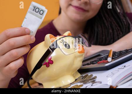 Ein Sparschwein wird von einem jungen Mädchen mit einer Papierbanknote im Nennwert von hundert polnischen Zlotys gefüttert. Ein zerrissenes Schweinebank aus Porzellan. Stockfoto