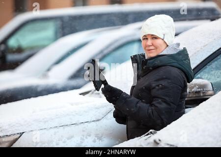 Die Fahrerin entfernt Schnee von der Vorderseite des verschneiten Autos, die Frau schaut auf die Kamera Stockfoto