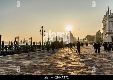 Venedig, Italien-Januar 28,2022. Menschen zu Fuß auf dem Damm, genießen sonnigen Wintertag, Kirche, Kanal und Gondeln im Hintergrund. Venezianischen Stadt Lebensstil. Stockfoto