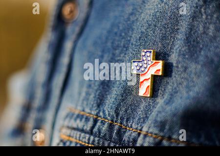 Die Nahaufnahme des Christian Cross Pin mit amerikanischen Fahnenfarben ist auf der blauen Jeansjacke fixiert. Patriotismus und religiöse Rechte Konzept Stockfoto