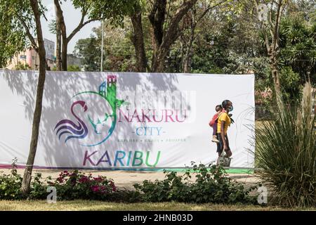 Nakuru, Kenia. 15th. Februar 2022. Eine Mutter, die ein Baby auf dem Rücken trägt, geht am Banner der Stadt Nakuru im Nyayo Garden im zentralen Geschäftsviertel vorbei.die Gemeinde Nakuru wurde vom Präsidenten Uhuru Kenyatta zum Stadtstatus hochgestuft, nachdem sie alle gesetzlichen Anforderungen erfüllt hatte.Sie wird nach Nairobi, Mombasa und Kisumu zur vierten Stadt. Kredit: SOPA Images Limited/Alamy Live Nachrichten Stockfoto