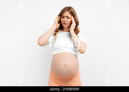 Porträt einer schönen jungen Schwangeren, die unter Kopfschmerzen auf weißem Hintergrund leidet. Kopfschmerzen aufgrund von Stress, Migräne. Stockfoto