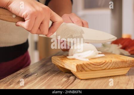 Person, die weiße Zwiebel mit einem Messer auf einem Holztisch in seiner Küche bequem von zu Hause aus schneidet Stockfoto