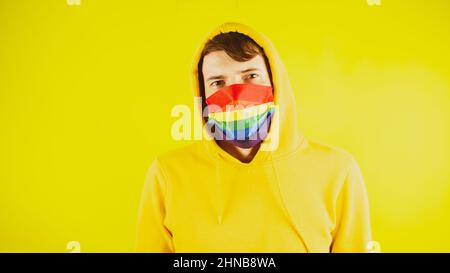 Mann in Maske lgbt im Studio. Junger Mann in gelbem Sweatshirt und Maske, der vor gelbem Hintergrund auf die Kamera blickt. Stockfoto