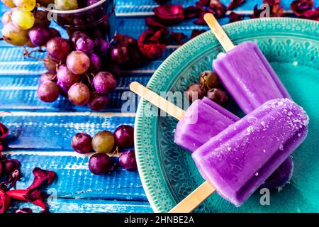 Ein Traubeneis im Lollipop-Stil auf einem Stock auf einem Vintage-Teller auf einem hellblauen Holztisch. Konzept der natürlichen und gesunden Ernährung. Copyspace. Stockfoto
