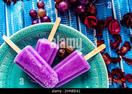 Traubeneis im Lollipop-Stil auf einem Stock auf einem Vintage-Teller auf einem hellblauen Holztisch. Konzept der natürlichen und gesunden Ernährung. Copyspace. Stockfoto