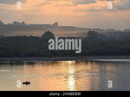 Landschaft mit einem Boot in den Morgenstrahlen. Morgenhimmel mit goldenen und düsteren Wolken über dem Ufer eines bewaldeten Flusses im Morgennebel. Die Wasserreferenz Stockfoto