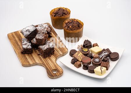 Brownies auf einem Holzbrett, Schokolade-Muffins und verschiedene Pralinen auf einem weißen Teller und weißem Hintergrund. Stockfoto