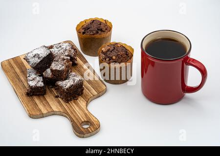Brownies auf Holzbrett, Schokolade-Muffins, verschiedene Pralinen auf einem weißen Teller, rote Kaffeetasse und weißer Hintergrund Stockfoto