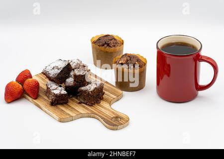 Brownies auf Holzbrett, Schokolade-Muffins, verschiedene Pralinen auf einem weißen Teller, rote Kaffeetasse, Erdbeeren und weißer Hintergrund Stockfoto