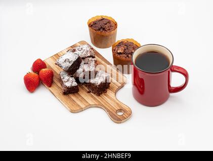 Brownies auf Holzbrett, Schokolade-Muffins, verschiedene Pralinen auf einem weißen Teller, rote Kaffeetasse, Erdbeeren und weißer Hintergrund Stockfoto