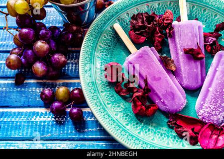 Ein Traubeneis im Lollipop-Stil auf einem Stock auf einem Vintage-Teller auf einem hellblauen Holztisch. Konzept der natürlichen und gesunden Ernährung. Copyspace. Stockfoto