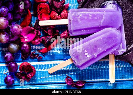 Three lollipop-style grape ice cream on a stick on a cast iron platter on a light blue wood table with some purple grapes on the side. Concept of natu Stockfoto