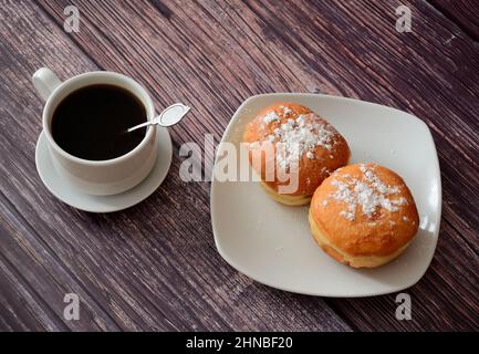 Eine Tasse frischen schwarzen Kaffee und ein Teller mit zwei Donuts in Puderzucker auf einem Holztisch. Draufsicht, flach liegend. Stockfoto