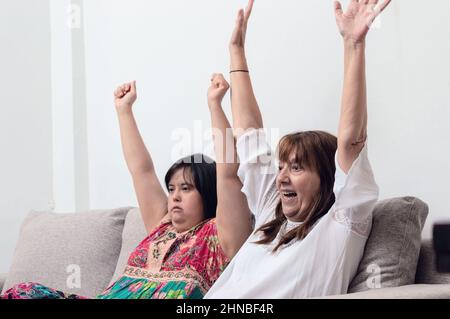 Mutter und Tochter mit Down-Syndrom, beim Fernsehen auf dem Sofa im Wohnzimmer sitzend, mit lächelnden, fröhlichen und feiernden Händen. Stockfoto