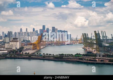 Der riesige verkehrsreichste Logistikhafen in Singapur, viele Krane, um Container zu bewegen, riesige Frachtschiffe im Hintergrund, Wolkenkratzer im Hintergrund Stockfoto