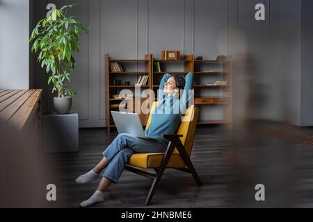 Fröhliche, lächelnde Freiberuflerin mit einem Laptop auf den Knien, die sich im Heimbüro mit den Händen hinter dem Kopf entspannt Stockfoto