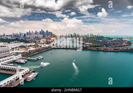 Der riesige verkehrsreichste Logistikhafen in Singapur, viele Kräne zum Transport von Containern, riesige Frachtschiffe im Hintergrund, Einkaufszentren und Wolkenkratzer Stockfoto