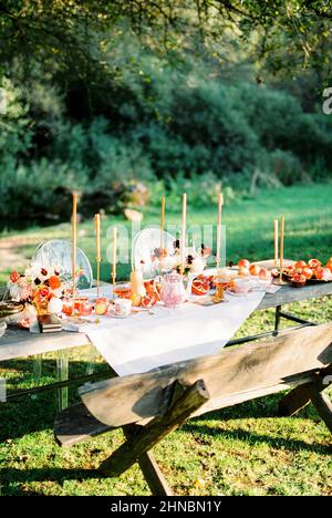 Gedeckte Tische mit Kerzen, Früchten und Getränken stehen neben einer Holzbank im Garten Stockfoto